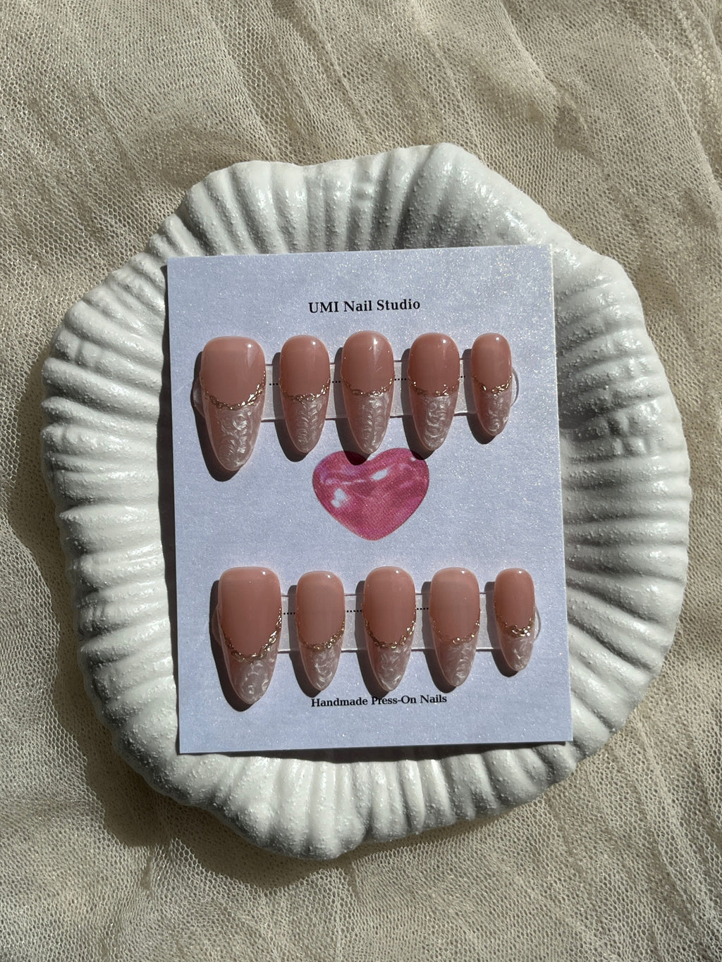 Nude pink press on nails with 3D white shell texture and gold chrome French tip line on a white shell display