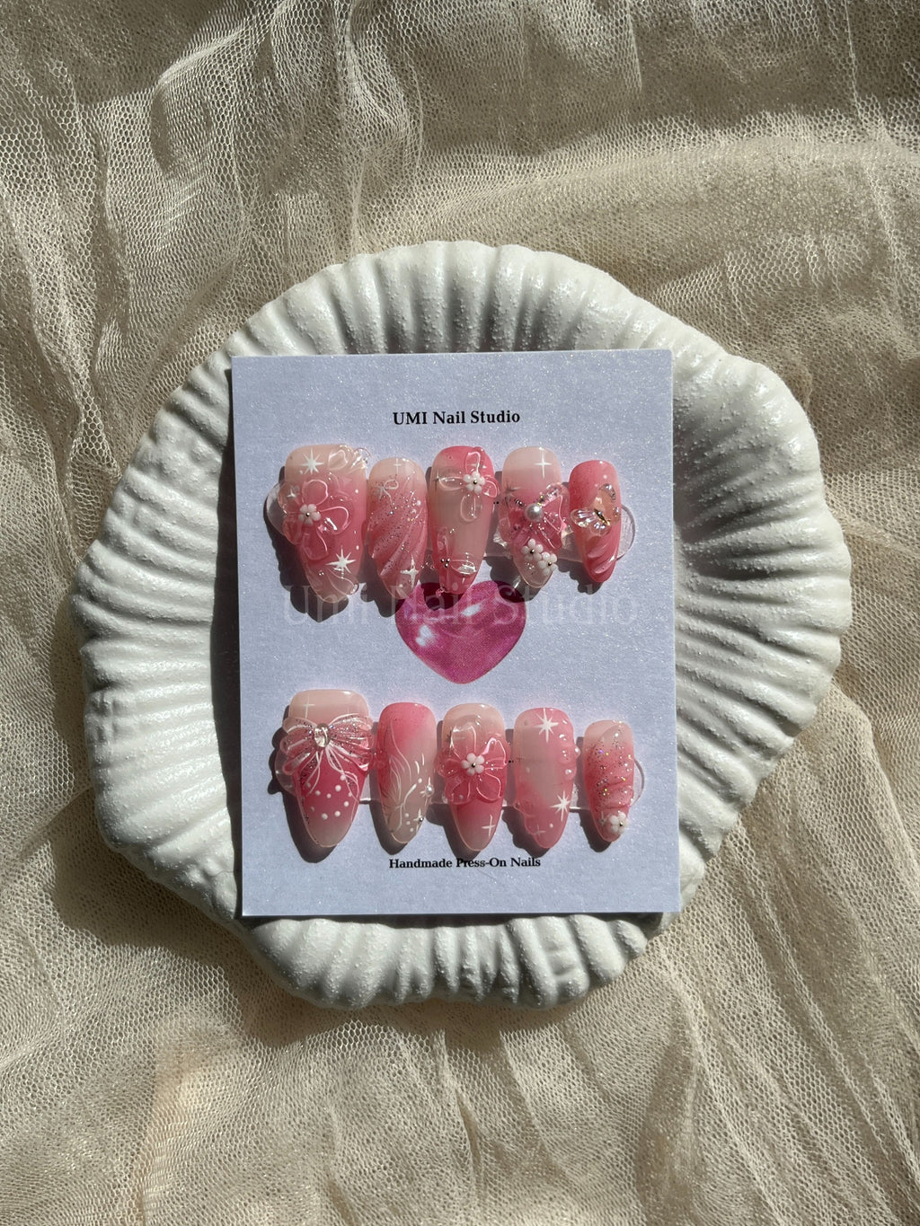 Handmade pink coquette press on nails with 3D flowers, pearls, and hand-painted white bows on a shell display.
